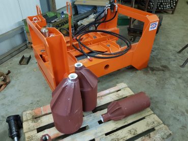 Pomarańczowy chwytak hydrauliczny ZRM oraz stalowe elementy wiertnicze na palecie w warsztacie.